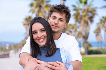 Fototapeta premium Young couple embrace on a palm lined beach promenade, boyfriend laughing as girlfriend wraps arms around his shoulders from behind, capturing joyful love, travel vibes, summer lifestyle, connection 