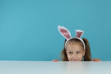 Curious little girl in bunny ears peeking over white table against light blue background, space for...
