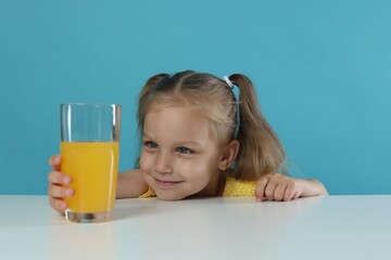 Cute little girl reaching for glass of orange juice at white table against light blue background