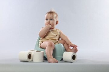 Cute little baby sitting on potty and rolls of toilet paper against light grey background