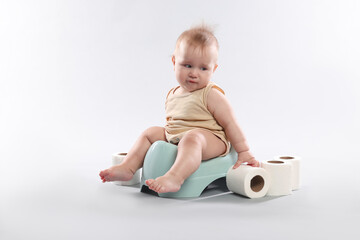 Cute little baby sitting on potty and rolls of toilet paper against light grey background