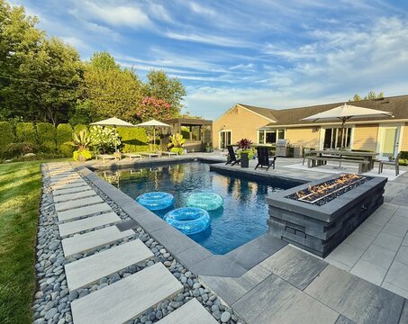 Beautiful, luxury, modern backyard lounge area. Umbrella, sofa, lounge chairs table poolside. Hotel resort feel. Seating and tanning sundeck by gray stone pavers. Fire pit and waterfall. High end. 