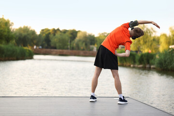 Sportsman in headphones and armband doing morning exercise on pier near river, back view. Space for...