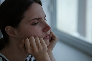 Sad young woman near window at home, closeup. Space for text
