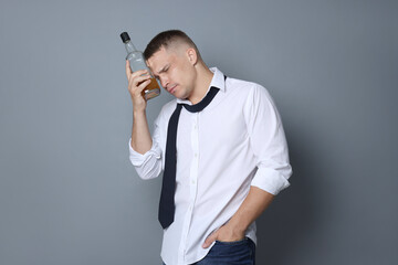 Man with bottle of whiskey suffering from hangover on grey background