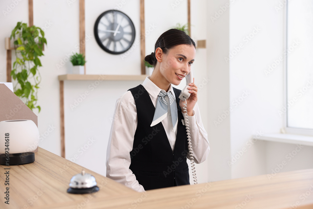 Poster smiling receptionist talking with client by phone at reception desk in hotel