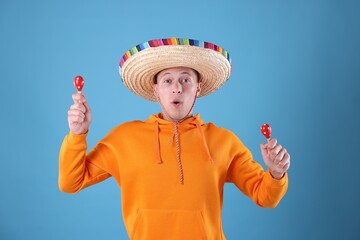 Emotional man in sombrero hat playing maracas on light blue background