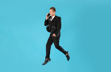 Businessman with folder jumping while talking on smartphone against light blue background