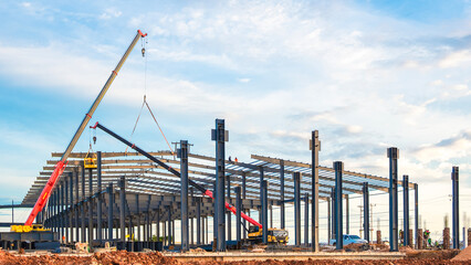 Large factory industrial building framework with workers and crane trucks are lifting metal roof beam to installing on top of warehouse structure in construction site against blue sky background
