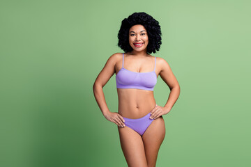 Charming young woman posing in lavender lingerie against a green background, embracing natural...