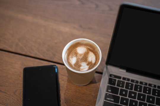 Close-up of Hot coffee latte with latte art milk foam in cup made of paper smartphone and laptop computer on wood desk office desk in coffee shop at the cafe,during business work concept