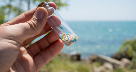 Highlighting the crisis of microplastic pollution: A hand holds a sample jar containing tiny microplastic fragments collected from a shoreline, showcasing the reality of water contamination.