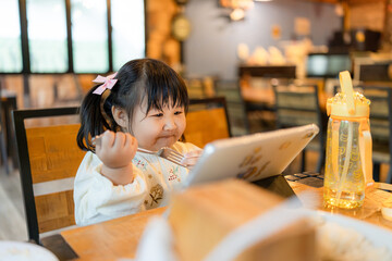 A little daughter sits next to her mother eating at a table in a restaurant and looks at a tablet screen,A cheerful girl is enjoying cartoons on a tablet with her mother in a restaurant.