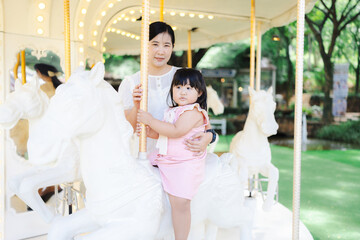 Mother and daughter are happily riding the carousel in the amusement park. Asian mother and cute...