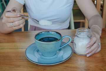 Young Asian woman is sitting at a table while using a spoon to add sugar to her coffee.