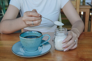Young Asian woman is sitting at a table while using a spoon to add sugar to her coffee.