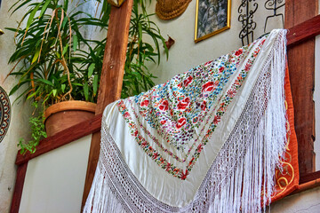 White embroidered shawl with colorful floral patterns and long fringe draped over a wooden railing,...