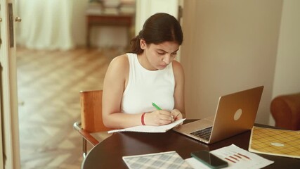 A dedicated student diligently writing notes at her desk using a laptop in a comfortable and cozy indoor setting - Powered by Adobe