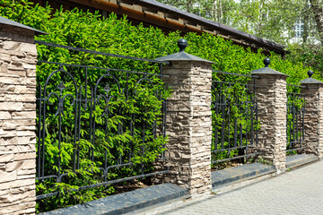 Elegant stone and wrought iron fence bordering a lush green hedge on a sunny day in a peaceful...