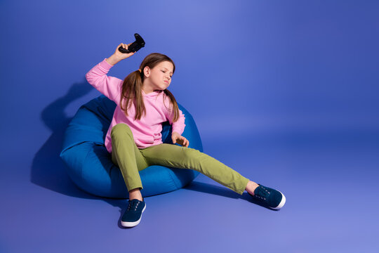 Preteen girl in bright casual outfit holding gaming controller on blue background, expressing emotions during leisure activity