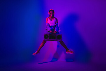 Stylish young woman holding a boombox in a vibrant violet and neon-lit room, expressing fashionable energy while dancing
