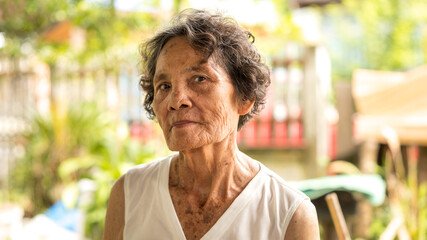 Elderly woman with curly hair stands outside in a lush garden during the afternoon light, showcasing wisdom and character © Happy Photo