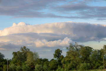 ​A view of the horizon, where massive, layered clouds hang over the green treetops. The calm light that breaks through the cloud formations creates a sense of majesty and the power of nature, 