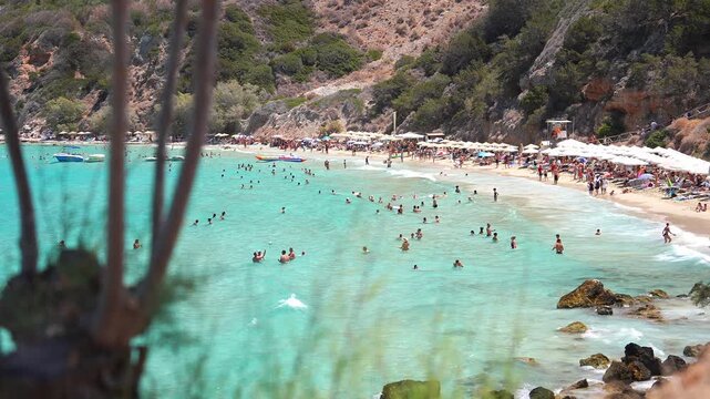 Turquoise waters meet a sandy beach bordered by cliffs and greenery in Crete, Greece. People swim, relax under umbrellas, and boats float near the shore.