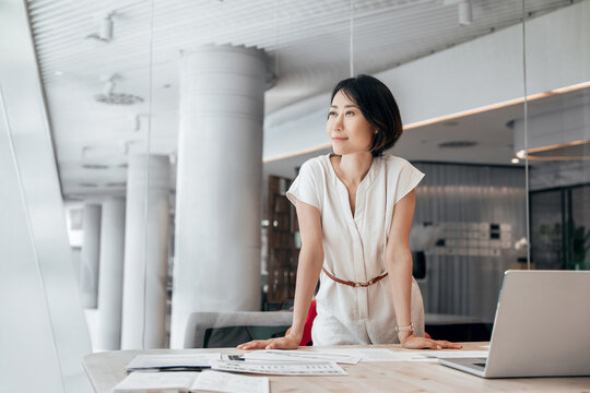 Thoughtful asian professional financial analyst specialist business lady working on laptop pc. Middle eastern woman investor dreaming standing standing at computer in modern office. Copy space