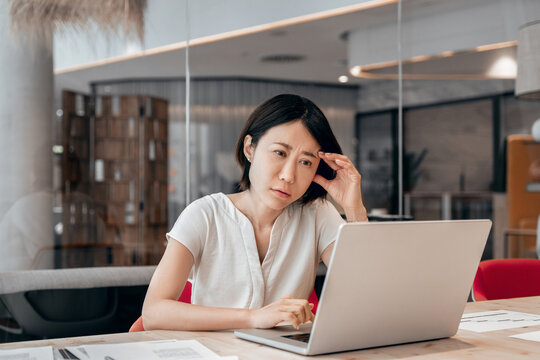 Focused young businesswoman looking at laptop pc computer screen having headache, migraine. Asian business woman holding hand near temples, feeling stressed and tired sitting at workplace in office