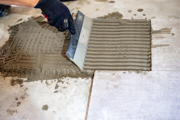 A construction worker applies adhesive mortar with a notched trowel before laying ceramic tiles on...