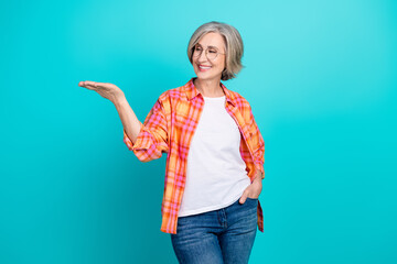 Smiling elderly woman posing casually in checkered shirt with hand gesture on teal background