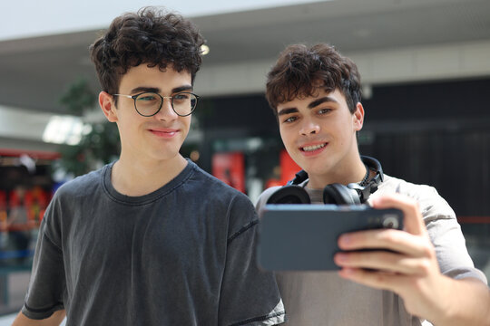 Two cheerful teenage twin brothers standing together and smiling while looking at a smartphone during a video call or live stream indoors. Casual urban youth lifestyle , friendship and family bonding
