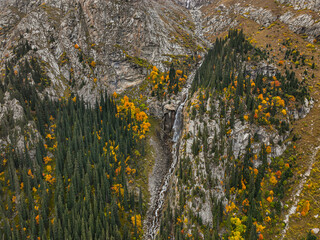 A detailed drone shot of a waterfall on a rugged rock face, framed by a mix of evergreen pines and...
