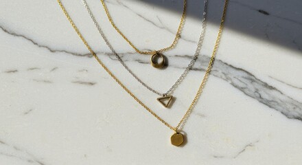 Three delicate gold and silver necklaces with geometric pendants lay on a marble surface.