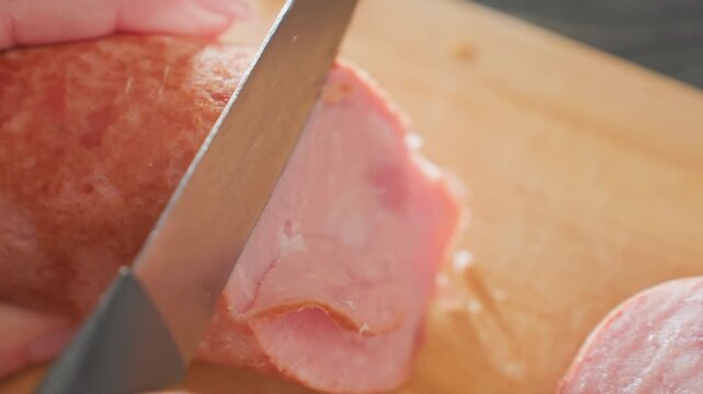close up hands slicing pink hotdog on wooden board using sharp kitchen knife on kitchen counter, showing texture of processed meat under soft lighting during indoor meal preparation