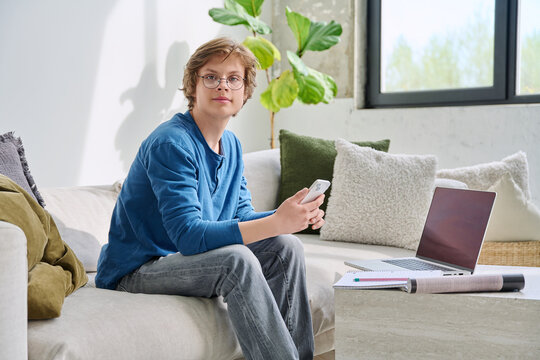 Teenager guy student studying sitting at home on sofa with smartphone laptop textbook