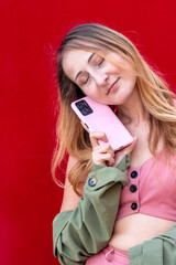 Young woman holding smartphone near face with closed eyes on red background