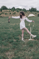 A curly-haired brunette woman in a short white cutout dress stands in a grassy meadow, holding a...