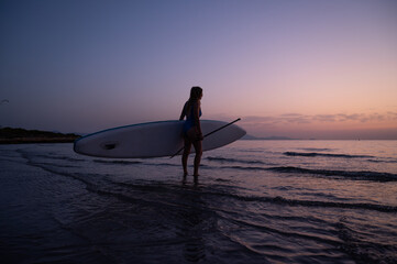 A woman is standing on the beach holding a surfboard. The sky is a beautiful mix of orange and...
