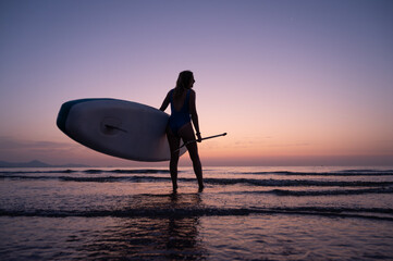 A woman is standing on the beach holding a surfboard. The sky is a beautiful mix of orange and...