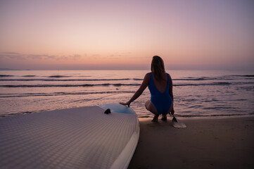 A woman is sitting on the beach with a surfboard in front of her. The sky is a beautiful mix of...