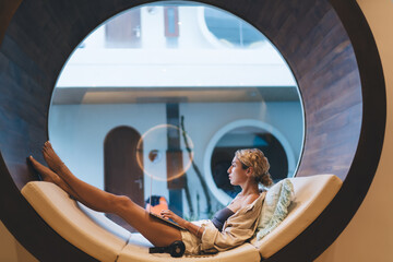 Focused woman types on laptop inside circular window nook, headphones nearby. Evoking futuristic workspace, tech immersion, and fluid transition between leisure and productivity zones.