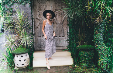 Stylish barefoot woman in summer dress and hat walking past artistic carved door, merging fashion...