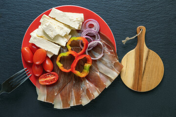 Balkan cuisine. Slices of prsut: dry-cured ham,  and local white cheese on red plate. Dark table,...