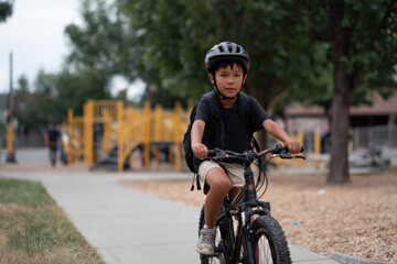Young boy riding bicycle wearing helmet and backpack near playground