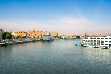 Luxury Nile cruise ships navigating through Esna Lock waterway with city buildings and dock infrastructure along riverbank in Egypt. (