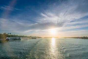 The sun sets over the Nile River, creating a golden reflection on the water's surface. A cruise ship is visible on the river with the lush green banks of Luxor in the distance.