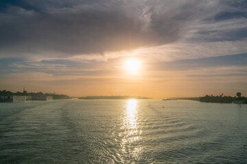 Naklejka premium Warm sunset light reflects on the Nile River near Esna Lock, Egypt, with boats, buildings, and trees lining the riverbanks.