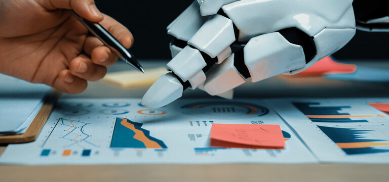 A human hand and a robotic hand work together on business charts, analyzing data trends and showcasing the fusion of technology and human creativity in the workplace.Noogenesis